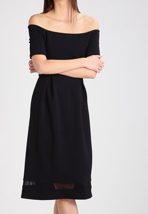 Femme portant une robe noire épaules dénudées à manches courtes, bras croisés, sur un fond clair uni.