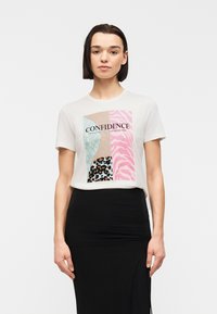 Γυναίκα με κοντά σκούρα μαλλιά, ντυμένη με λευκό t-shirt με πολύχρωμο ζωικό μοτίβο και την επιγραφή «CONFIDENCE» και μαύρη φούστα με ψηλή μέση.