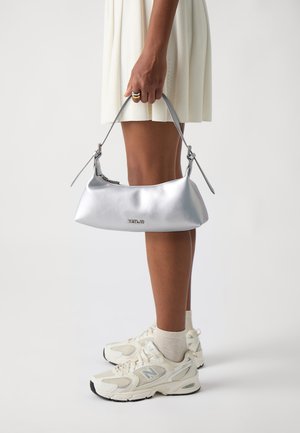 Silberne metallische Handtasche mit einem eleganten, geschwungenen Design und verstellbarem Riemen, gehalten von einer Person in einem hellen Kleid, kombiniert mit weißen Sneakers.