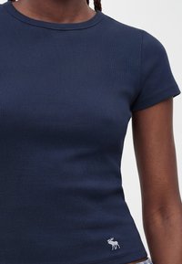 Κοντινό πλάνο ενός ατόμου που φοράει μπλε σκούρο ριμπ μπλουζάκι με κοντά μανίκια και μικρό λευκό λογότυπο ελάφι κοντά στο τελείωμα στην αριστερή πλευρά.
