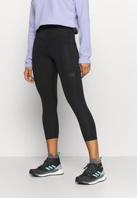 Osoba ubrana w czarne, krótkie legginsy z logo The North Face, fioletową bluzkę z długim rękawem oraz czarne i turkusowe sneakersy Adidas, stojąca.