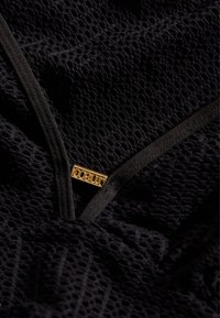Tissu en maille noire avec un motif texturé et une bordure noire lisse, arborant une étiquette logo dorée intégrée dans le design.