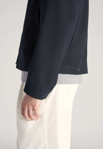 Granatowa kurtka o miękkiej fakturze, z zapięciem na zamek, bocznymi kieszeniami i lekko luźnym kroju, noszona na szarym t-shircie i białych spodniach.