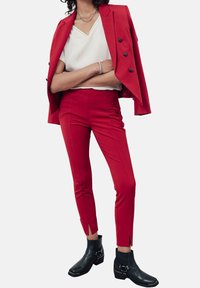 Personne portant une veste rouge drapée sur les épaules, un haut blanc en V, un pantalon rouge ajusté, des bottines noires à la cheville, un collier en chaîne argenté et un bracelet.