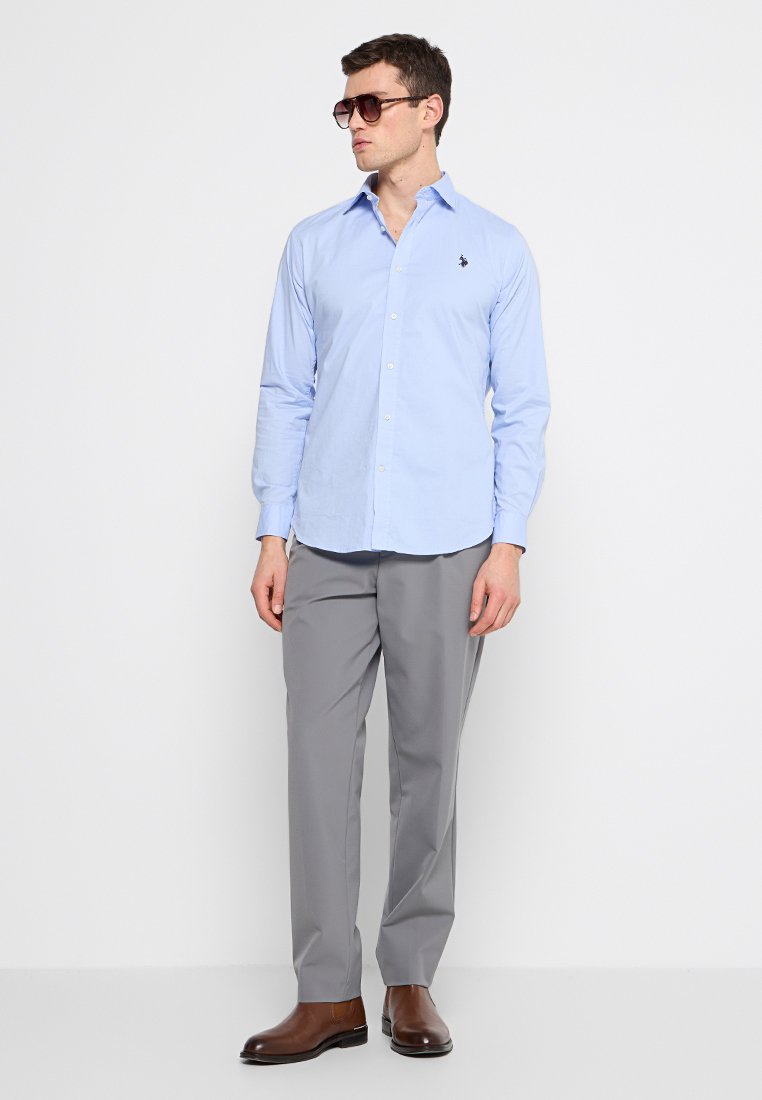 Uomo che indossa una camicia azzurra chiara con bottoni, pantaloni grigi, scarpe in pelle marrone e occhiali da sole scuri, in piedi davanti a uno sfondo bianco.
