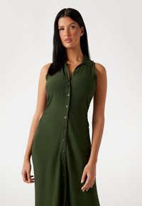 Khaki vihreä hihaton mekko, jossa on nappikiinnitys edessä, kaulus ja muototikkinen siluetti, valmistettu sileästä ja kevyestä kankaasta.
