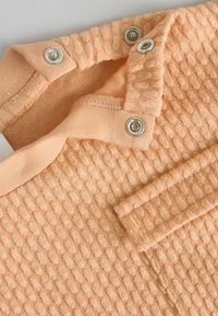 Pfirsichfarbener Strickstoff mit strukturiertem Muster, mit Druckknöpfen entlang des Halsausschnitts und einem umgeschlagenen Kragen. Weich und leicht.