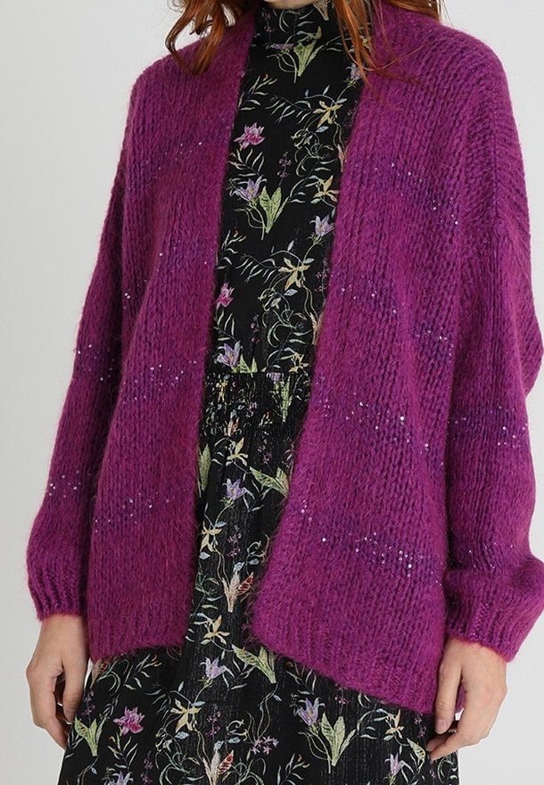 Femme portant un cardigan tricoté ample de couleur violet foncé sur une robe noire à fleurs avec des manches longues et un col haut.