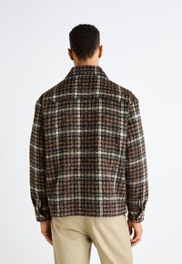 Brązowa kratkowana kurtka z kołnierzem i zapinanymi mankietami. Teksturowany materiał z ciemnymi i jasnymi kwadratami. Widok z tyłu pokazuje krótki fason.