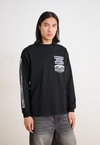 Black long-sleeve t-shirt featuring front and sleeve graphics labeled "MANASTASH SPORTS COMMITTEE." Fabric appears smooth, with a relaxed fit.