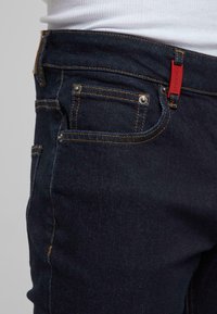 Gros plan d'un jean denim bleu foncé avec des coutures orange, montrant une poche avant et une petite étiquette rouge sur la boucle de ceinture, porté avec une chemise blanche côtelée.