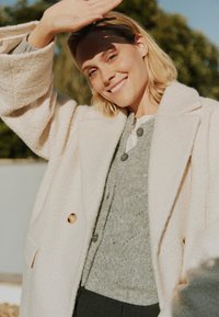 Beige, strukturierter Mantel mit großen Knöpfen, über einem hellgrauen Strickpullover mit subtilen Mustern und Perlenakzenten getragen, kombiniert mit dunklen Hosen.