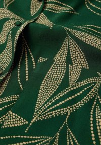 Grüner Stoff mit einem Blattmuster in goldenen Punkten. Das Design umfasst fließende Formen und eine glatte Textur mit leichtem Glanz.