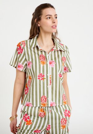 Kobieta ubrana w zielono-biały pasiasty krótki rękaw i szorty z różowo-pomarańczowymi nadrukami kwiatów i ananasa.
