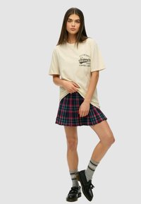 Camiseta de algodón beige con logo, combinada con una falda corta plisada a cuadros en tonos azul marino, verde y rojo. Zapatos negros y calcetines grises con texto.