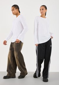 Deux personnes avec les cheveux tressés portant des chemises blanches à manches longues ; l'une en jean large marron, l'autre en pantalon de survêtement noir Adidas avec des rayures.
