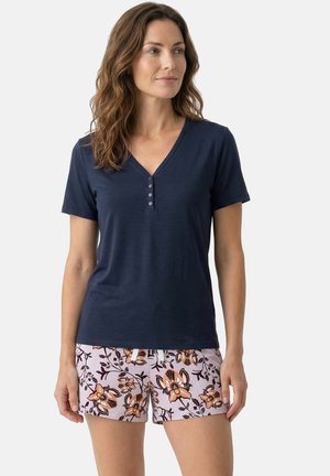 Vrouw met golvend bruin haar die een marineblauw T-shirt met korte mouwen en V-hals draagt en lichtroze korte broek met bloemenpatroon, staand tegen een effen achtergrond.