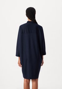 Laivansinis ylipitkä mekko, jossa on 3/4-hihat, kaulus ja saumaa yläselän kohdalla, valmistettu sileästä kankaasta, jossa on minimaalinen rakenne.