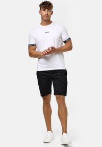 T-shirt blanc avec logo et manches noires, associé à un short noir. Modèle portant des baskets blanches. Fond neutre.