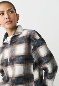 Fleece quarter-zip trøje med et ternet mønster i hvide, grå og brune nuancer. Har en høj krave og en blød tekstur.