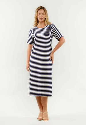 Naine kannab põlveni ulatuvat lühikeste varrukatega tumesinise-valge triibulise kleidiga ja beeže sandaale, seisab tavalise valge tausta ees.