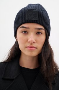 Black ribbed knit beanie with a textured black patch displaying a geometric logo. Worn with a black coat and crew-neck sweater.