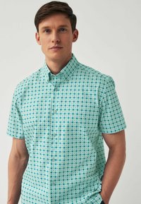 Kurzarmhemd in Türkis mit einem geometrischen weißen Muster. Mit Button-down-Kragen und weichem Stoff. Verfügt über eine Brusttasche und dezente Knöpfe.