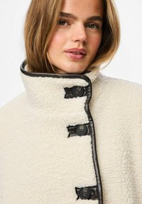 Abrigo de forro polar color crema con un cuello alto, ribetes de cuero oscuro y tres grandes botones negros. Textura suave con un acabado liso.