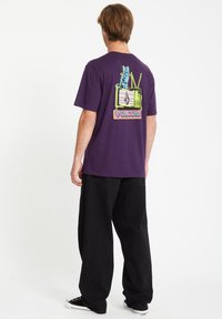 Lila Baumwoll-T-Shirt mit einem grafischen Druck einer Katze, die auf einem Retro-Fernseher sitzt. Hat einen lässigen Schnitt und kurze Ärmel, kombiniert mit schwarzen Hosen.