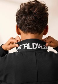 Schwarze Jacke mit hohem Kragen, der weiße Schriftzüge aufweist. Der Stoff wirkt strukturiert und glatt. Die Haare sind kurz und lockig.