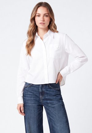 Kvinde med langt bølget hår iført en hvid skjorte med knapper og blå jeans, stående med den ene hånd på hoften mod en hvid baggrund.