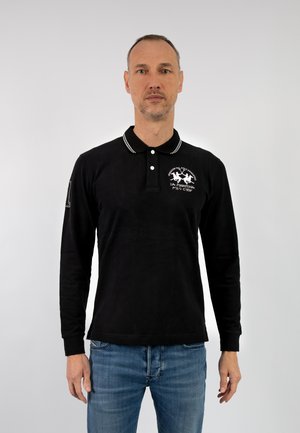 Polo shirt nero a maniche lunghe con patta a due bottoni, strisce bianche sul colletto e grafica ricamata sul petto. Indossato con jeans blu.