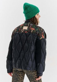 Padded jacket z czarnym pikowanym korpusem i kwiatowym wzorem na ramionach. Posiada naszywkę oraz akcenty z czarnego polaru. Noszona zielona czapka.