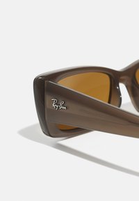 Ochelari de soare din plastic maro cu lentile galbene. Prezintă un design geometric și logo-ul Ray-Ban pe brățară. Textură netedă, fără modele.