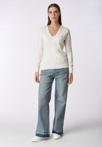 Maglione bianco a V realizzato in tessuto morbido, abbinato a jeans larghi blu chiaro e sneakers bianche. Dettaglio del logo ben visibile sul maglione.