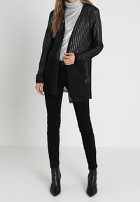 Černý průsvitný kardigan s metalickými pruhy, otevřený vpředu, ve spojení se šedým rolákem a černými úzkými džínami, doplněný černými kotníkovými botami.