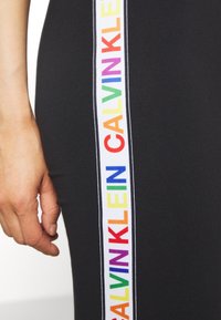 Černé legíny s vertikálním bílým pruhem a textem "CALVIN KLEIN" v duhových barvách, zobrazené na ruce poblíž pasu.
