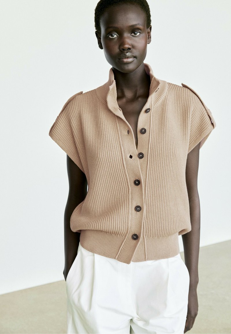 Woman wearing a beige sleeveless knitted buttoned vest with a high collar and white loose-fitting trousers, standing against a plain background.