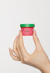 Rosa beholder med grønn lokk, merket "Velsmakende Vannmelon Leppepeeling." Har svarte frømønstre, 20 ml kapasitet. Holdt av en hånd.