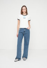 Λευκό κοντομάνικο μπλουζάκι με μαύρο περίγραμμα και μπλε λογότυπο Converse. Συνδυάζεται με ανοιχτό μπλε τζιν παντελόνι φαρδιάς γραμμής και ασημί παπούτσια τύπου slip-on.