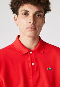 Polo rojo con tejido texturizado, tres botones y un logo verde de Lacoste en el lado izquierdo del pecho. Cuello corto y diseño ajustado.