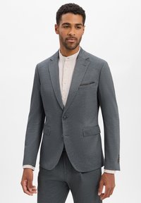 Grijze textuurpak met een enkel-breasted blazer, notch-lapels en twee voorzakken. Gedragen over een lichtgekleurde overhemd met kraag.