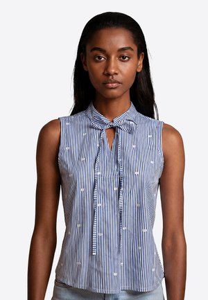 Donna che indossa una camicia senza maniche a righe verticali blu e bianche con piccoli fiocchi ricamati e un fiocco annodato al collo.