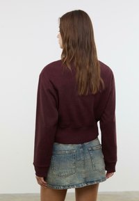 Donna con lunghi capelli castani che indossa un maglione bordeaux e una gonna di denim blu sbiadito, in piedi con la schiena rivolta alla macchina fotografica contro un muro neutro.