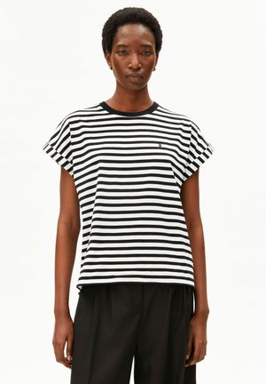 Vrouw draagt zwart-wit horizontaal gestreept T-shirt met korte mouwen en zwarte broek, staand tegen een effen witte achtergrond.