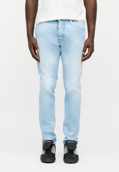 Homme portant un jean en denim bleu clair, une chemise blanche et des baskets noires à rayures blanches, debout devant un fond uni.