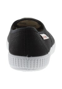 Baskets slip-on noires en toile avec une semelle en caoutchouc blanche et un talon texturé. Présente une petite étiquette logo multicolore sur le côté.