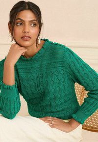 Pull vert en maille avec un motif en losanges texturé, doté d'un col et de poignets froncés, ainsi que de boutons décoratifs à l'épaule.