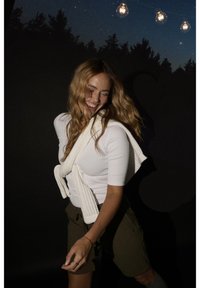 Femme souriante aux longs cheveux ondulés, portant une chemise blanche et un pull posé sur les épaules, se tient devant un ciel nocturne sombre avec des lumières suspendues.