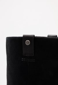 Black suede boot featuring a leather strap with a circular metal logo accent. Smooth texture with visible stitching details.